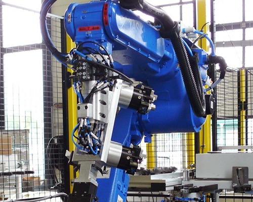 Industrieaufnahme eines blauen, mehrachsigen Knickarmroboters innerhalb einer Sicherheitsumhausung mit gelben Schutzpfosten und Metallgitter. Am Roboterhandgelenk ist ein komplexes, mehrteiliges Greif‑ und Handhabungssystem aus gefrästem Aluminium montiert, bestehend aus mehreren parallel arbeitenden Greifmodulen. Sichtbar sind pneumatische Leitungen, Ventilblöcke, Sensoren sowie elektrische Anschlusskabel, die präzise entlang der Greiferstruktur geführt sind. Das Greifsystem ist für das positionsgenaue Halten, Ausrichten oder Montieren von Werkstücken ausgelegt und an einer automatisierten Fertigungs‑ oder Montageanlage eingesetzt. Im Hintergrund sind weitere Maschinenkomponenten, Werkstückträger und industrielle Infrastruktur erkennbar, was auf eine robotergestützte Serienproduktion in einer Fertigungshalle hinweist.