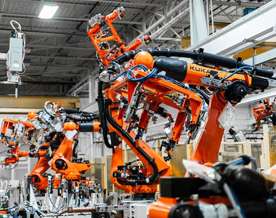 Mehrere orangefarbene KUKA‑Industrieroboter mit Greifarmen arbeiten entlang einer automatisierten Fertigungslinie in einer Industriehalle und handhaben Werkstücke im Rahmen der Gießereiautomation. Im Hintergrund sind weitere Roboter, Anlagen und die Hallenkonstruktion zu sehen.