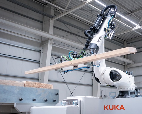 Industrieroboterarm in einer Fabrikhalle, der einen langen Holzträger horizontal greift und positioniert, montiert auf einer automatisierten Produktionsanlage. Auf dem weißen Roboterarm ist deutlich der Schriftzug „KUKA“ zu sehen, im Hintergrund befinden sich Stahlträger, Beleuchtung und weitere Maschinenelemente der Industrieumgebung.