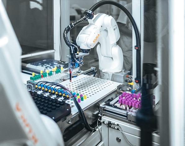 Ein weißer KUKA‑Industrieroboterarm arbeitet in einer geschlossenen Laboranlage und handhabt automatisiert Probenröhrchen mit Blutproben, die in farblich gekennzeichneten Haltern auf einer Arbeitsplattform angeordnet sind. Umgeben ist das System von Greiftechnik, Schläuchen und weiteren Komponenten der Laborautomation.
