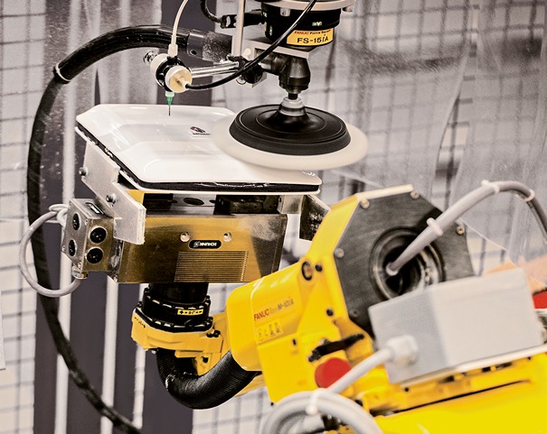 Ein gelber FANUC‑Industrieroboter ist mit einem rotierenden Polierwerkzeug ausgestattet und bearbeitet automatisiert die Oberfläche eines eingespannten Werkstücks innerhalb einer Industrieanlage. Schläuche, Sensoren und Schutzvorrichtungen für die Polishing‑Oberflächentechnik sind sichtbar.